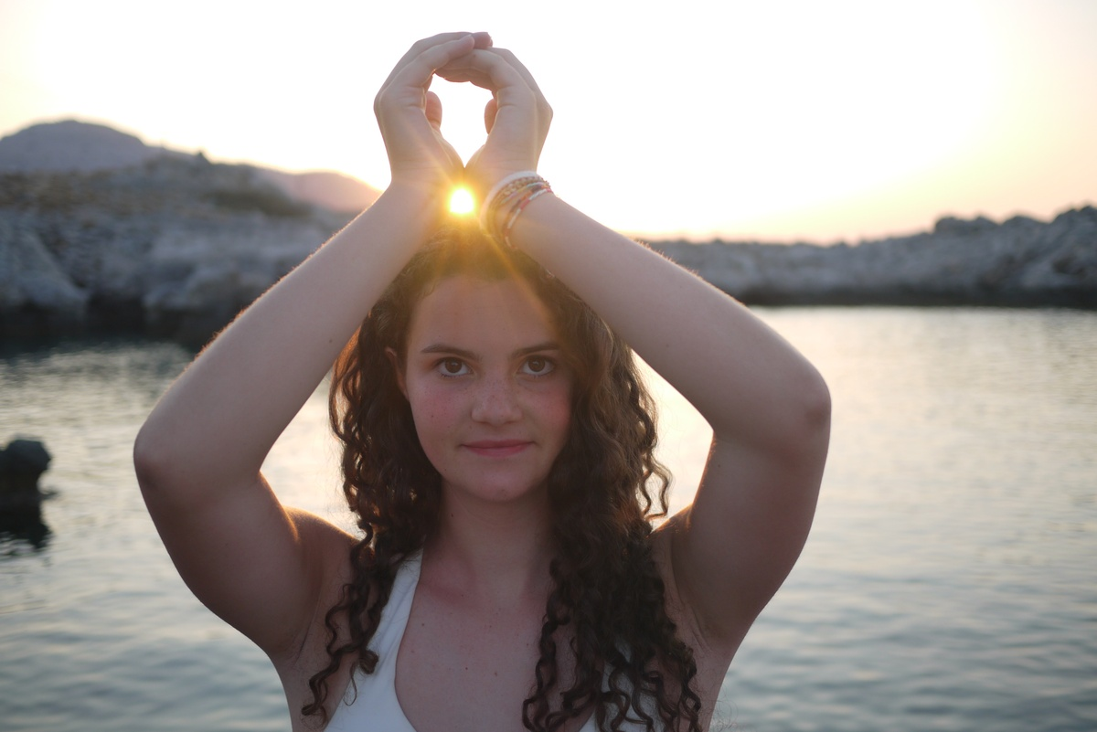 Mujer con las manos levantadas formando un círculo con el sol al atardecer junto al agua